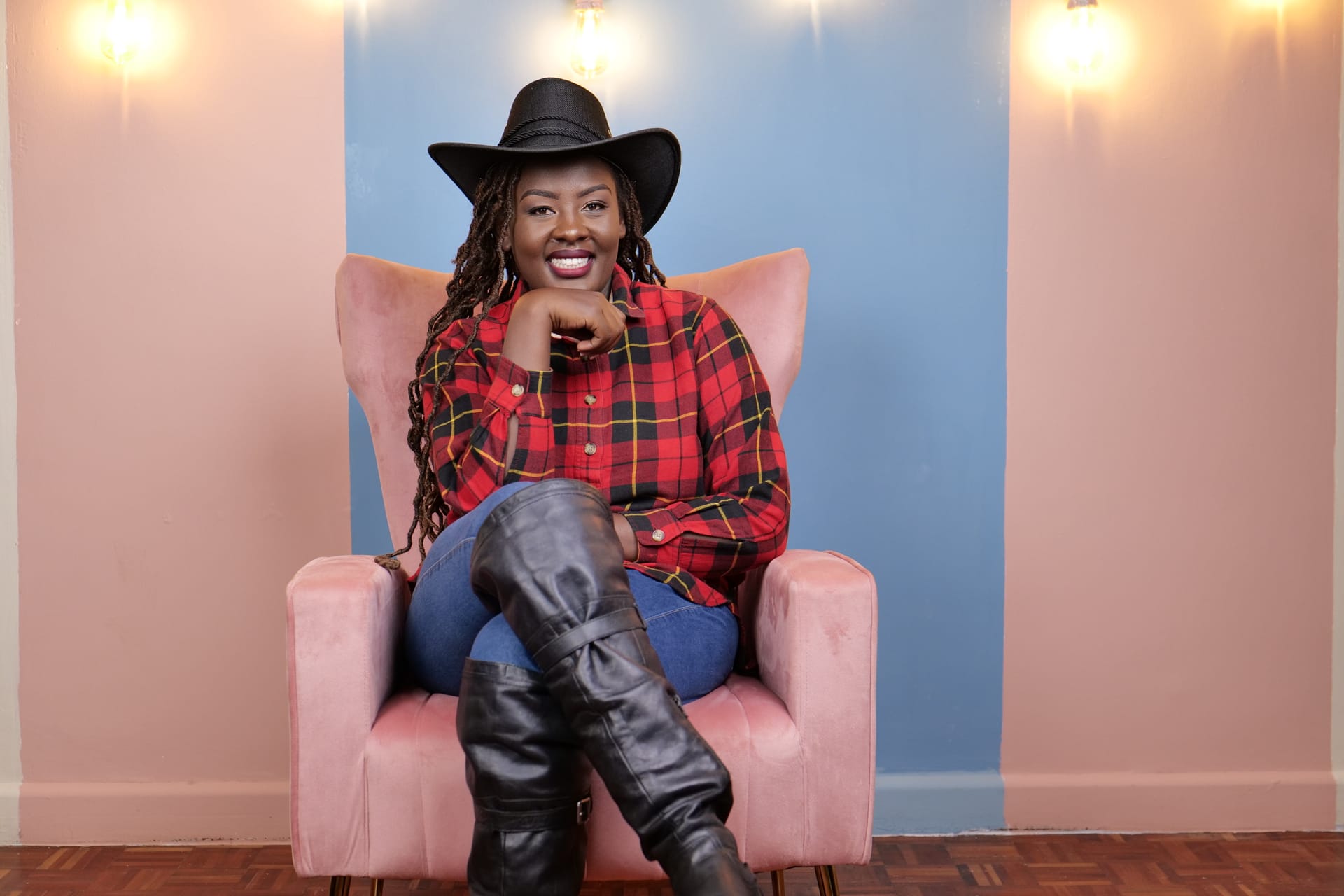 Serah in a black wide-brimmed hat and red plaid shirt, seated in a pink armchair.