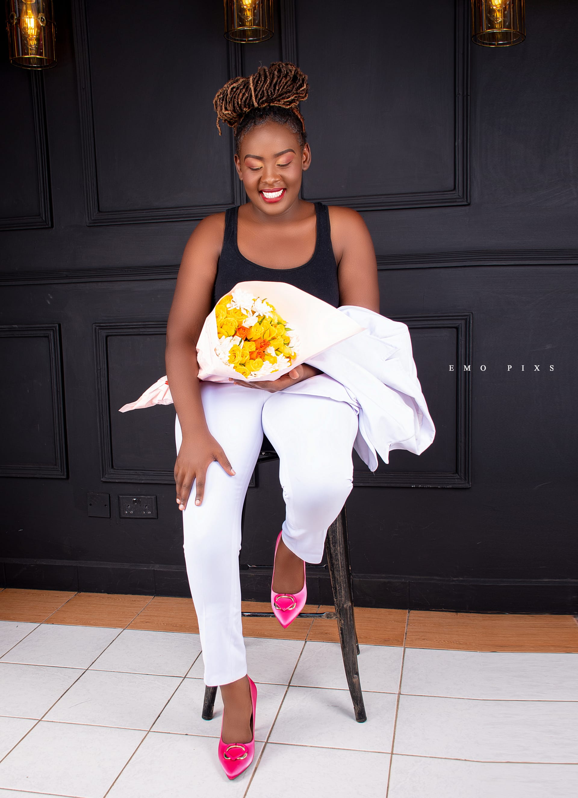 Serah Ke seated against a dark panelled wall, laughing, holding a bouquet of yellow flowers.